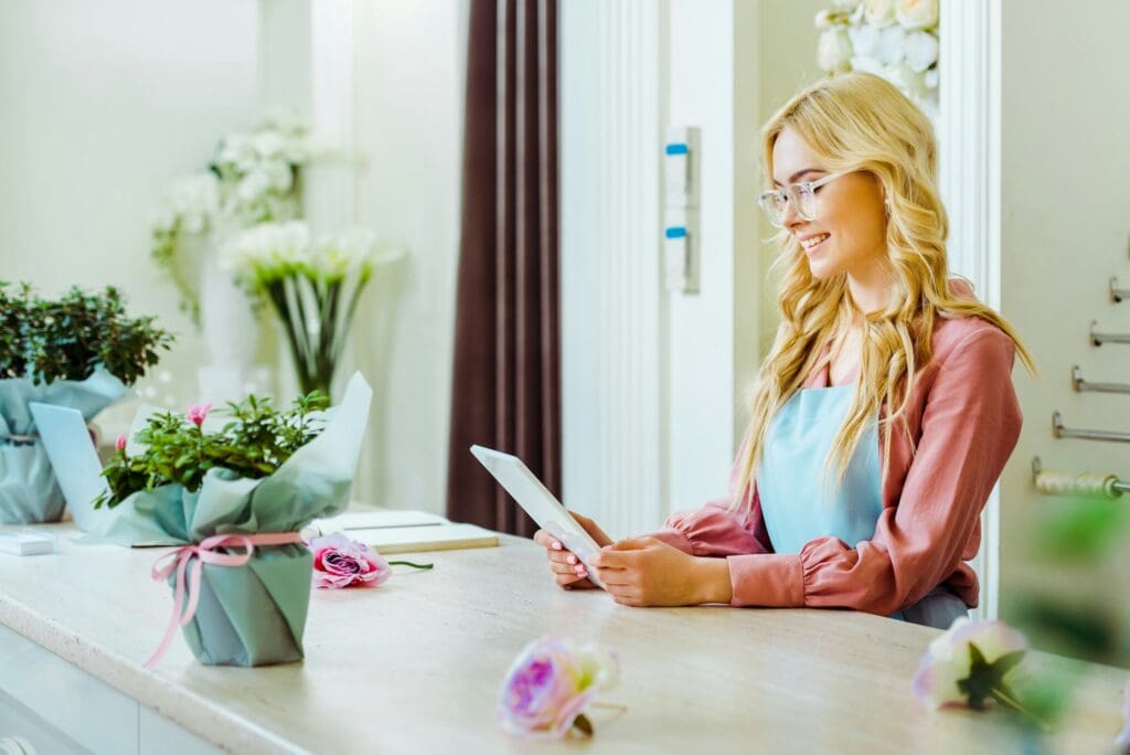 beautiful smiling female flower shop owner in glasses using digital tablet at counter
